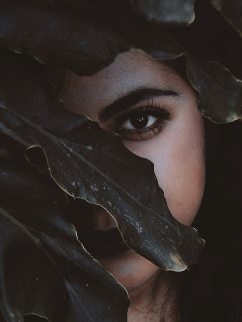 A woman's eye peers through lush dark leaves, creating a mysterious and captivating atmosphere. - Credits: pexels