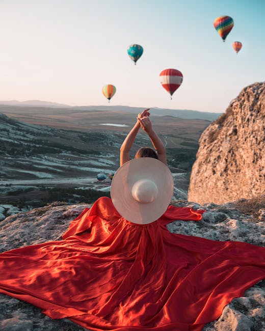 A woman in a red dress and hat enjoys a scenic view with hot air balloons over a rocky landscape. - Credits: pexels.com